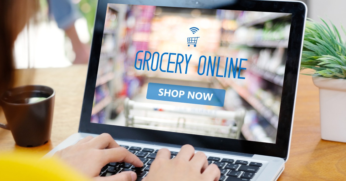 A woman wearing a yellow shirt sits at a desk with a laptop, focused on ordering groceries on a website.